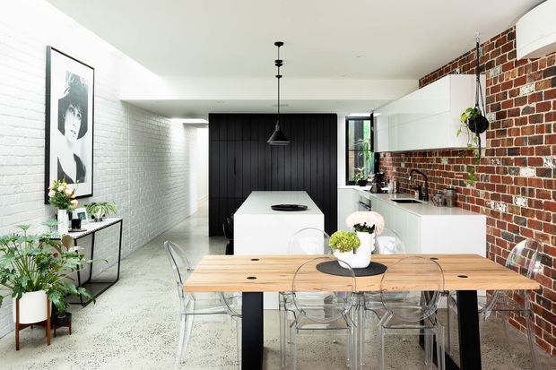 A bright contemporary kitchen and dining area with a long timber dining table and transparent chairs in the foreground. The kitchen features a white island with a black panelled feature wall, white cabinetry, exposed red brickwork, pale terrazzo-style flooring, and pendant lighting. Indoor plants and framed artwork add soft decorative accents.