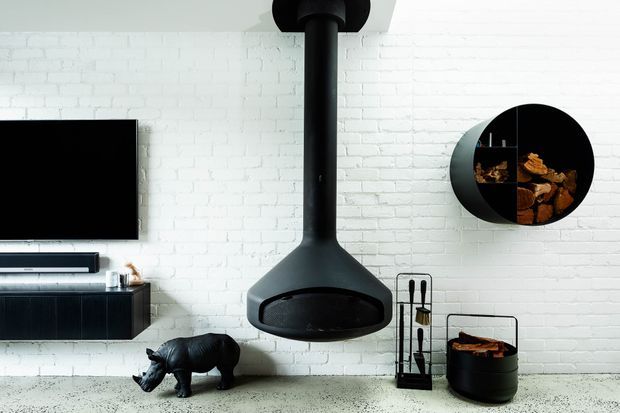 A modern living room with a white-painted brick feature wall, a suspended black wood-burning fireplace, wall-mounted black media cabinetry and television, and a round black shelving unit displaying firewood. A black sculptural animal and firewood storage add decorative detail. The space has a restrained monochromatic palette with industrial elements.