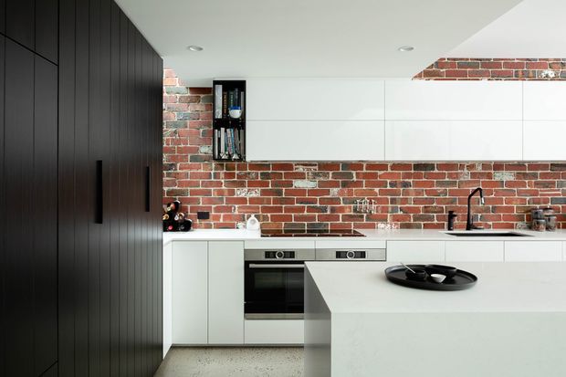 Contemporary kitchen with a large white island, integrated white cabinetry, black vertical pantry cabinetry, exposed red brick splashback and wall finish, and modern integrated appliances. A black sink tap and recessed ceiling lights provide contrasting accents.