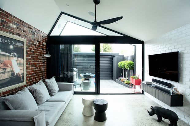 Contemporary living room with a pale sectional sofa, black sculptural coffee tables, wall-mounted television and large black-framed glazed doors opening to a small courtyard. The room combines exposed red brick, white-painted brick, a polished light concrete floor, a dramatic ceiling fan and abundant daylight.