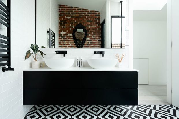 Contemporary bathroom with a large black floating double vanity, two white vessel basins, wall-mounted black tapware, mirrored wall, white subway-tile walls, and a bold black-and-white geometric floor. A red brick feature wall and bright adjacent hallway are reflected in the mirror.