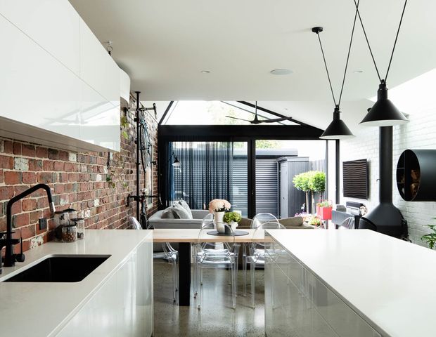 Bright open-plan kitchen and dining/living space with glossy white cabinetry and island benches, a white benchtop, exposed red brick wall, polished concrete floor, black-framed glazing, and a glazed roof or clerestory bringing in natural light. Black pendant lights and a sculptural black fireplace provide strong contrast, while a courtyard is visible beyond the dining area.