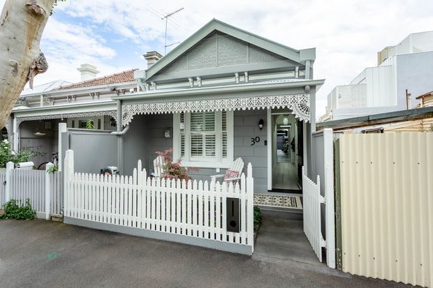 Street-facing view of a compact heritage-style single-storey cottage with a gabled roof, grey weatherboard cladding, ornate lacework verandah detailing, white picket fencing, and a small front garden. A contemporary building is visible alongside it.