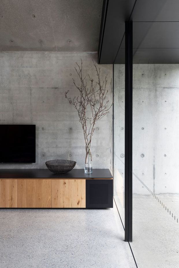 Minimalist living room interior featuring a raw exposed concrete wall and ceiling, a low built-in timber and dark joinery media console, wall-mounted television, and full-height black-framed glazing or an internal glass partition. A simple vase with bare branches and a sculptural wire bowl add restrained natural and decorative accents. The space is monochromatic, tactile, and strongly influenced by industrial architecture.