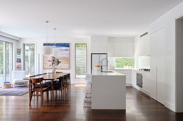 Bright open-plan kitchen and dining area with white floor-to-ceiling cabinetry, a large central island, integrated stainless-steel appliances, dark timber flooring, and a timber dining table with mid-century style chairs. Large windows and glazed doors bring in abundant natural light, while artwork and shelving add visual interest.