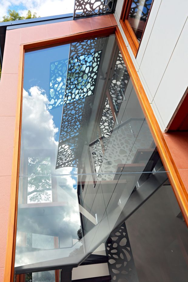Upward-looking architectural detail of a contemporary building facade with extensive reflective glazing, warm timber window frames, pink-toned cladding panels, and white wall panels. Decorative perforated metal screens with an organic leaf-like pattern create layered shadows and reflections across the glass. Interior stair or circulation elements are visible through the glazing, while sky and trees reflect on the windows.