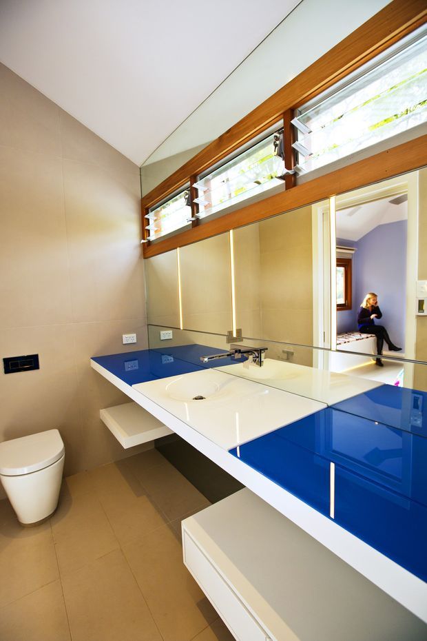 A contemporary bathroom with a floating white vanity, integrated white basin, vivid glossy blue benchtop and cabinetry, large mirrored wall, beige tiled walls and floor, and warm timber-framed high-level louvre windows. A toilet is visible at left, while the mirror reflects an adjoining bedroom area.