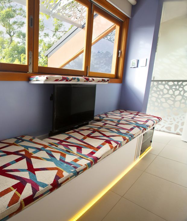 Compact, brightly lit interior nook with purple walls, warm timber-framed windows, pale tiled flooring, and a long built-in upholstered bench or daybed. The bench has bold multicoloured geometric fabric upholstery and integrated warm LED lighting beneath it, with a television set into the wall-side joinery. Decorative patterned privacy screens are visible outside and on the glazed door or partition.