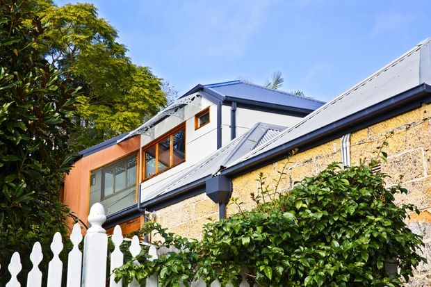 Exterior view of a renovated multi-level residence combining an older pale stone or masonry structure with a contemporary upper extension. The building has pitched corrugated metal roofs, dark gutters and downpipes, large timber-framed glazed openings, and warm timber cladding. A white decorative picket fence and dense greenery occupy the foreground.