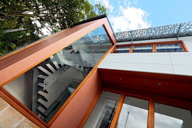 Upward-looking architectural exterior view of a contemporary multi-level building, showing a glazed stairwell with a floating or open-riser staircase, warm timber-framed glazing and soffits, pale horizontal wall cladding, and an ornate perforated metal privacy screen along the upper terrace or roof edge. Mature greenery and sky surround the building.