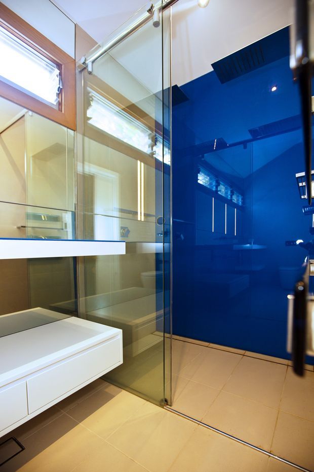 Modern bathroom with a frameless glass shower enclosure, vivid glossy blue feature wall, pale beige floor and wall tiles, white floating vanity, and timber-framed windows. The mirrored and glazed surfaces create strong reflections and make the compact room feel brighter.