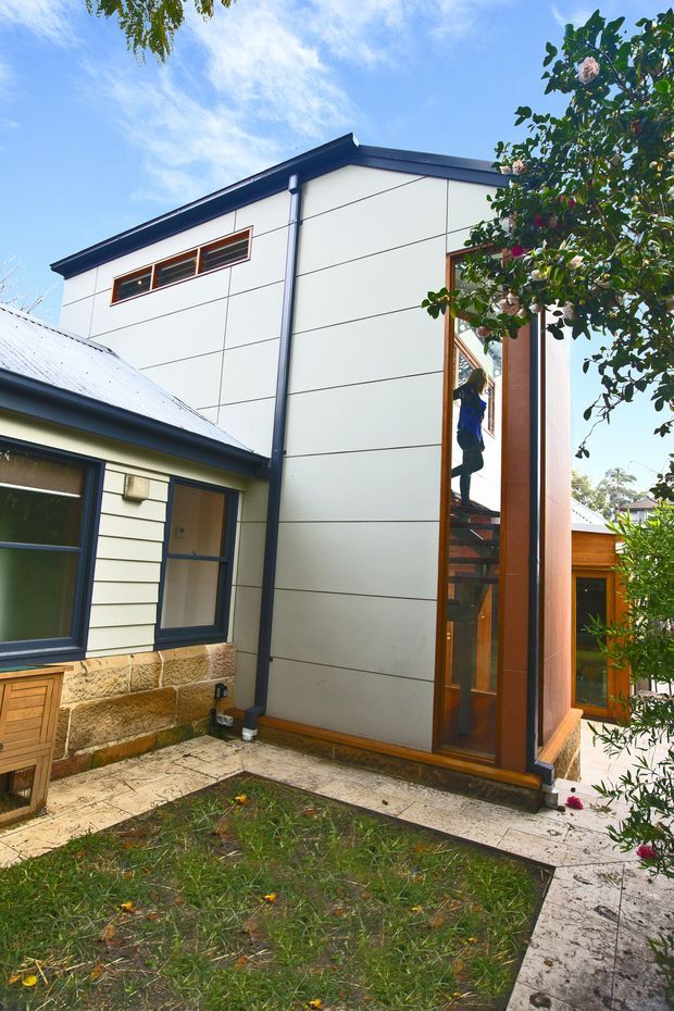 Exterior view of a contemporary residential extension attached to an older weatherboard house. The new upper-level volume has smooth white panel cladding with expressed horizontal and vertical joints, dark metal roof edging and downpipes, and a tall timber-framed glazed stairwell. The adjoining original structure features pale weatherboard cladding, a corrugated metal roof, dark blue window frames, and exposed stonework at the base. A small paved courtyard and lawn sit in front, with mature planting partially screening the building.