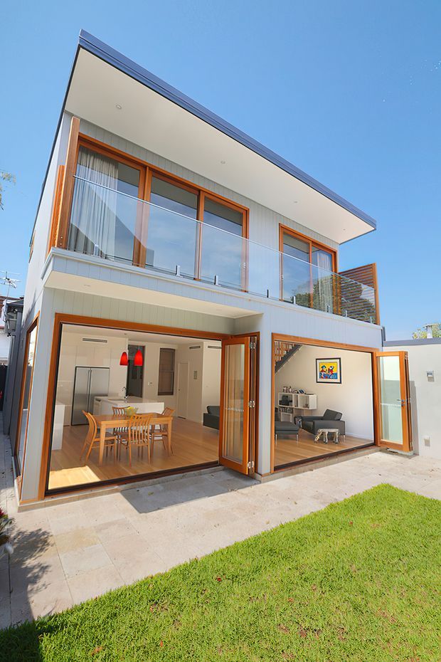 Exterior view of a contemporary two-storey residence with a broad flat roof, pale vertical cladding, warm timber-framed glazing, a glass-balustraded upper balcony, and large open bi-fold doors. The ground floor reveals a dining and kitchen area on the left and a living room on the right, opening onto a paved lawn. The facade is bright and minimalist with strong indoor-outdoor connection.