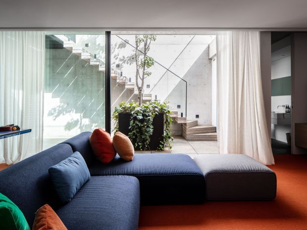 Contemporary living room with a large navy sectional sofa and ottoman on a vivid red-orange carpet. Floor-to-ceiling glazing with sheer curtains opens visually onto a sculptural concrete courtyard with steps, planting, and a small tree. The room has a restrained palette of dark blue, warm red, white, and concrete grey, with a partially visible bathroom or utility space beyond the opening at right.