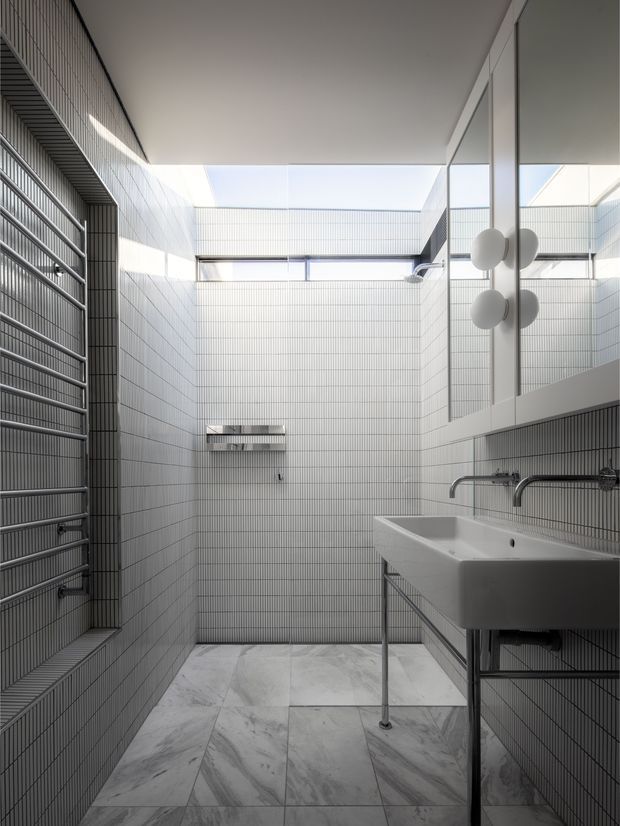 A compact, highly modern bathroom with full-height small-format white wall tiles, large pale marble-look floor tiles, an exposed chrome towel radiator on the left, and a wall-mounted industrial-style basin with open metal supports on the right. A large mirrored cabinet and spherical wall lights sit above the basin. High-level glazing and a bright skylit opening bring natural light into the space.