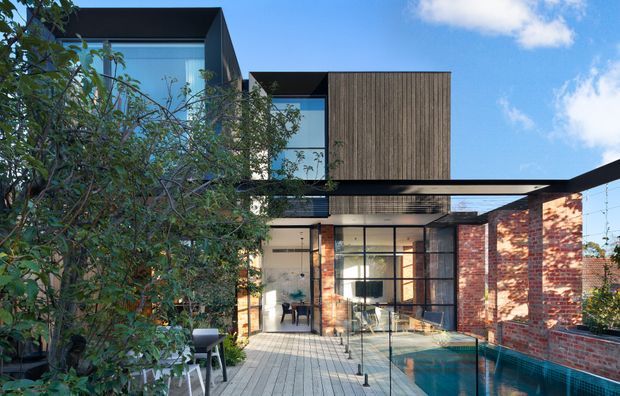 Contemporary multi-level residential facade with large glazed openings, dark flat roof forms, vertical timber cladding, exposed red brick walls, and a paved courtyard terrace beside a narrow swimming pool. Mature planting partially screens the structure, while a deep black-framed canopy projects over the outdoor living area.