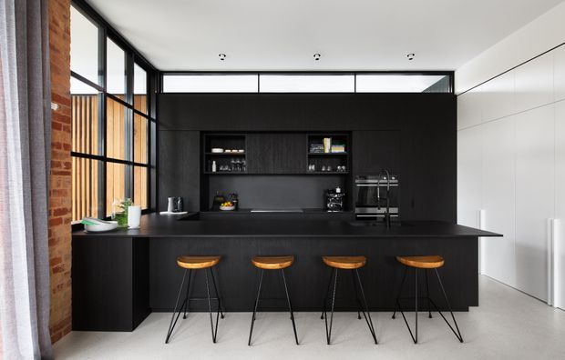 Contemporary kitchen with a long black island and four bar stools, dark built-in cabinetry, integrated shelving and appliances, white full-height cabinetry, exposed brick, timber screening, and extensive black-framed glazing beneath a high ceiling.