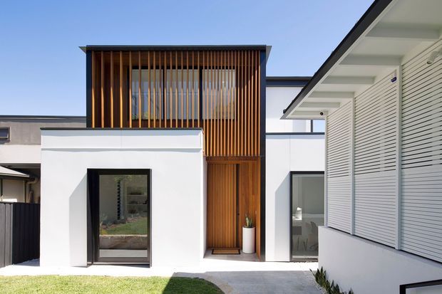 Bright contemporary residential exterior showing a white rendered building with a projecting upper volume screened by vertical timber battens, a timber-clad entry door, dark-framed glazing, and a neighbouring wing with horizontal louvred shutters. The low, clean-lined composition includes a paved path and small lawn in the foreground.