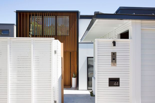 Contemporary residential facade viewed from the street, featuring a white horizontal-slat entry gate and fence, a dark flat roof, and a warm timber privacy screen with vertical battens around the upper-level glazing. The recessed entry is marked with a black number plaque reading 44 and a modern intercom. Clean geometric lines and contrasting white, charcoal, and timber finishes create a refined modern exterior.