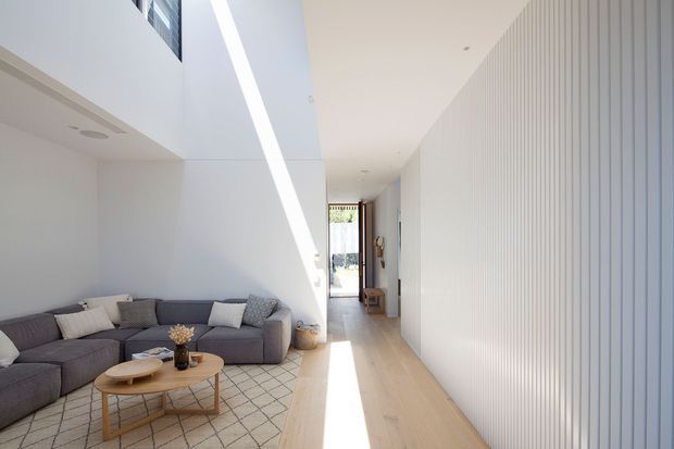 Bright contemporary open-plan living room and hallway with a low grey sectional sofa, round timber coffee table, pale timber flooring, white walls, and a large geometric-patterned rug. A dramatic diagonal shaft of sunlight falls across the double-height space, while vertical white wall panelling defines the right side and a glazed doorway leads toward the rear of the home.