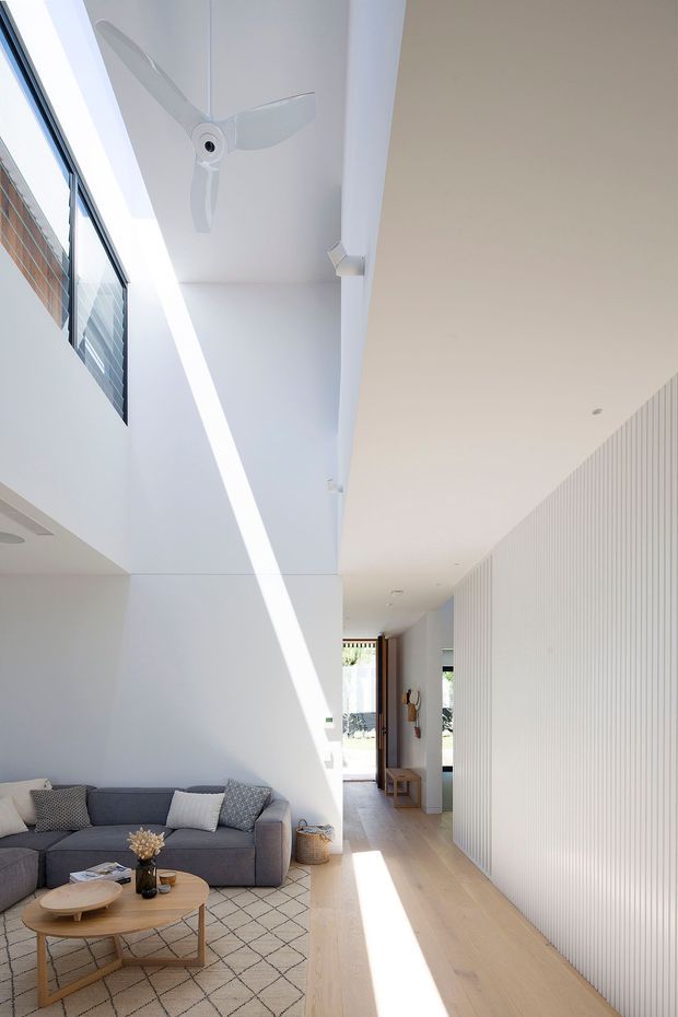 Bright, narrow contemporary living room and circulation space with a very high ceiling and strong linear daylight. The room features a grey sectional sofa, round timber coffee table, patterned rug, white walls, pale timber flooring, a white ceiling fan, high-level windows, and a full-height fluted or ribbed white wall along the hallway. A timber-framed glazed doorway is visible at the far end.