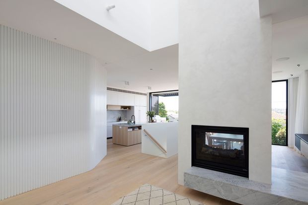 Bright contemporary open-plan interior featuring a minimalist white kitchen and island, pale timber flooring, a curved ribbed feature wall, and a central white fireplace with a stone hearth. Large windows and glazed doors provide abundant natural light and views to the outdoors.