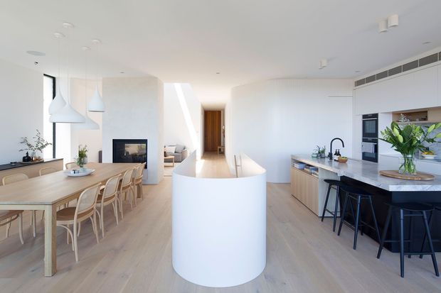 Bright open-plan contemporary kitchen and dining space with a sculptural curved white island, pale timber flooring, light wood cabinetry, white surfaces, integrated appliances, a long dining table, and minimalist pendant lighting. A living area and fireplace are visible beyond the dining zone.