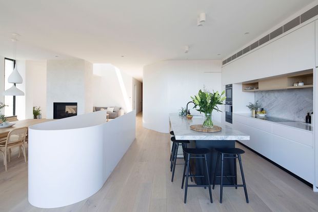 Bright open-plan contemporary kitchen and living area with a large white marble-look island, integrated white cabinetry, pale timber flooring, curved partition wall, and a fireplace visible in the adjoining living space. Black stools, pendant lights, indoor plants, and a vase of greenery add contrast and warmth.