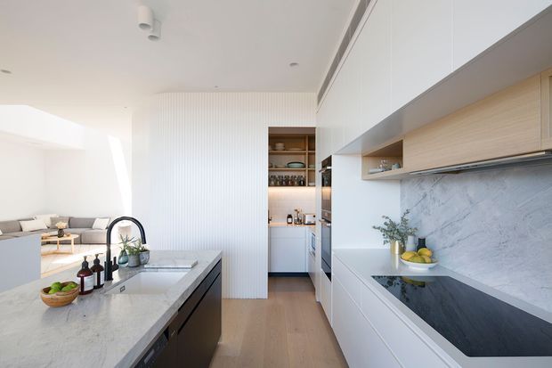 Bright, refined open-plan kitchen with white full-height cabinetry, pale timber joinery, a large stone island, and a dramatic veined stone splashback. The kitchen opens to a minimalist living area, with warm timber flooring, integrated appliances, open shelving, and abundant natural light.