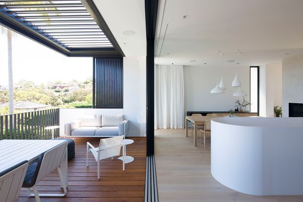 Bright, open-plan contemporary living and dining space opening onto a timber deck. Large retractable glazed doors and a louvred pergola create a strong indoor-outdoor connection, with a pale curved kitchen island, light timber flooring, white cabinetry and walls, sculptural pendant lights, and restrained modern furnishings. The deck overlooks a leafy suburban landscape.