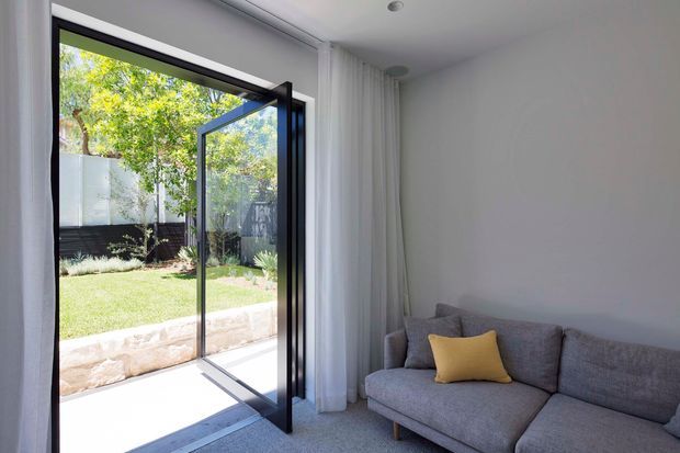Bright contemporary living room corner with a grey sectional sofa and yellow accent cushion, opening through a slim black-framed glazed door to a landscaped garden. White walls, sheer curtains, recessed ceiling lights, and pale carpet create a clean, airy interior with strong indoor-outdoor connection.