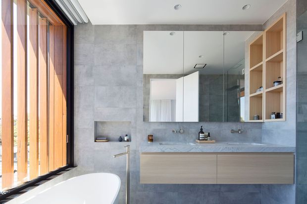 Contemporary bathroom with a freestanding white bathtub beside full-height timber-framed glazing and vertical timber screening. Grey stone-look wall and floor finishes surround a floating light timber vanity with a pale stone benchtop, twin basins and wall-mounted taps. Large mirrored cabinetry and open timber shelving occupy the main wall, creating a bright, refined space with natural textures and clean-lined joinery.