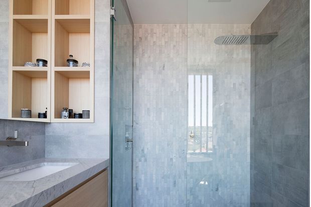 Contemporary bathroom featuring a walk-in glass shower with a large overhead rain shower, pale vertically patterned wall tiles, grey stone-look wall and benchtop surfaces, and light timber open shelving and vanity cabinetry. The room is bright and clean with a restrained neutral palette and natural wood accents.
