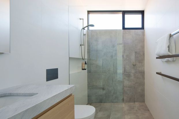 Bright contemporary bathroom with a walk-in shower enclosed by a clear glass screen, pale grey stone-look wall and floor tiles, white wall surfaces, and a floating timber vanity with a light stone benchtop. A wall-hung toilet, dark flush plate, chrome shower fittings, and open timber towel shelves are visible.
