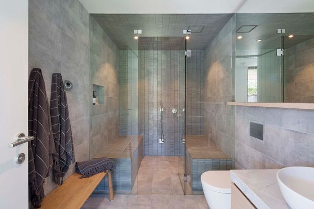 Contemporary bathroom with a spacious walk-in glass shower, full-height grey and taupe tiled walls, matching tiled ceiling, built-in tiled benches, timber seating, wall-mounted fixtures, a floating vanity with a white vessel basin, and a toilet. A large mirror enhances the sense of space and reflects the shower area.