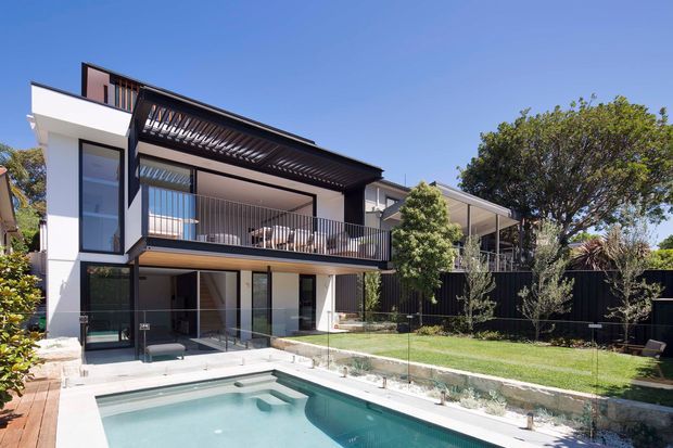 Contemporary multi-storey residential home with a crisp white and charcoal facade, extensive glazing, projecting upper-level balcony, and a dark pergola or louvred canopy. The rear yard includes a swimming pool, lawn, landscaped planting, and a covered outdoor area. The image is photographed from the backyard, emphasizing the building facade and pool setting.