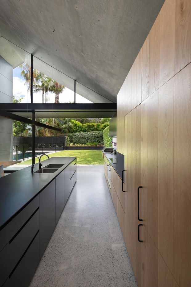 A narrow, modern kitchen opening directly onto a landscaped backyard and pool area. The space features a long dark kitchen island with an inset double sink and black tapware, extensive light timber wall cabinetry with black handles, dark lower cabinetry, a polished concrete ceiling, and a terrazzo-like concrete floor. Full-height glazing and a glazed roof connection bring in abundant daylight and frame the garden.