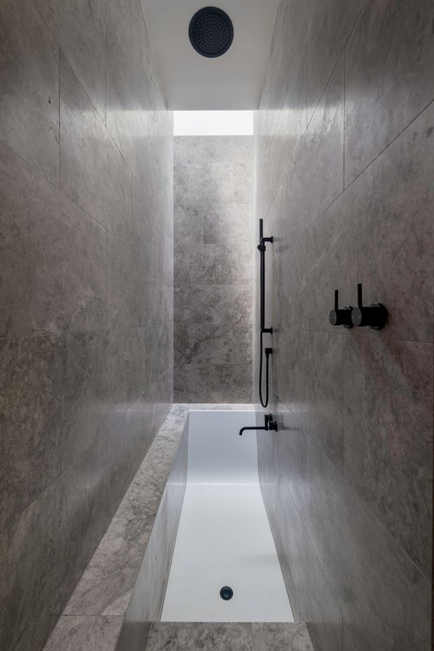 A narrow contemporary bathroom showing a full-size white freestanding bathtub beneath a ceiling-mounted shower, with a separate handheld shower rail and matte black controls. Large-format grey stone-look tiles cover the walls and floor, and a bright skylight or clerestory opening provides natural light at the end of the room.