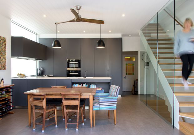 Open-plan kitchen and dining area with dark matte cabinetry, integrated appliances, a timber dining table and chairs, and colourful striped dining chairs. A glass-sided stair with timber treads rises beside the kitchen, while a timber-bladed ceiling fan, pendant lights, white panelled ceiling, and large glazing complete the bright contemporary interior.