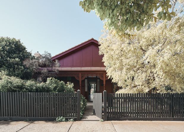 Front exterior view of a compact heritage-style house, partly screened by mature trees and a tall dark timber picket fence. The facade features red-brown brickwork, ornate timber detailing, a covered verandah with a corrugated roof, and a steep gabled upper section with deep red cladding. A narrow pedestrian gate and path lead to the recessed entry.