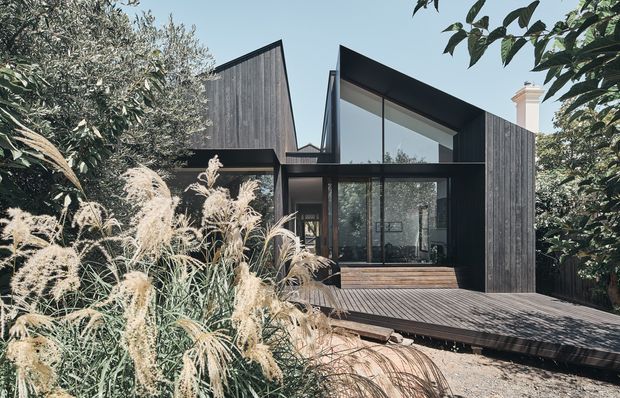 A contemporary dark-clad house set within dense garden landscaping. The compact composition features sharply angled roof forms, extensive floor-to-ceiling glazing, a timber deck and a central glazed living area, with ornamental grasses and mature trees screening the facade.
