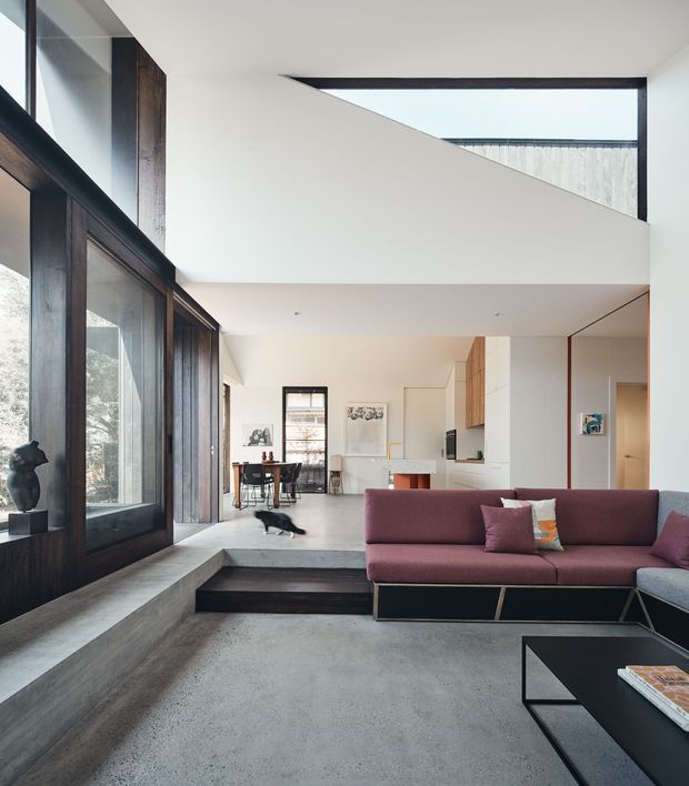 A refined open-plan living room with a burgundy sectional sofa, black coffee table, and a raised dining and kitchen area beyond. Full-height dark timber-framed glazing lines the left side, while a dramatic angular ceiling opening and double-height volume bring in abundant natural light. Concrete flooring and stepped levels create a strong contemporary architectural character.