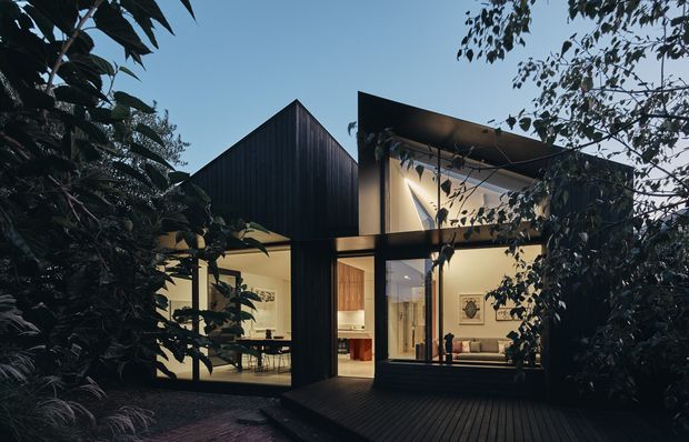 Dusk exterior view of a contemporary dark-clad residence set within dense garden planting. The house has strong angular forms, a prominent gabled volume, a flat-roofed glazed living area, extensive floor-to-ceiling windows, and a timber deck leading into warmly lit interiors.
