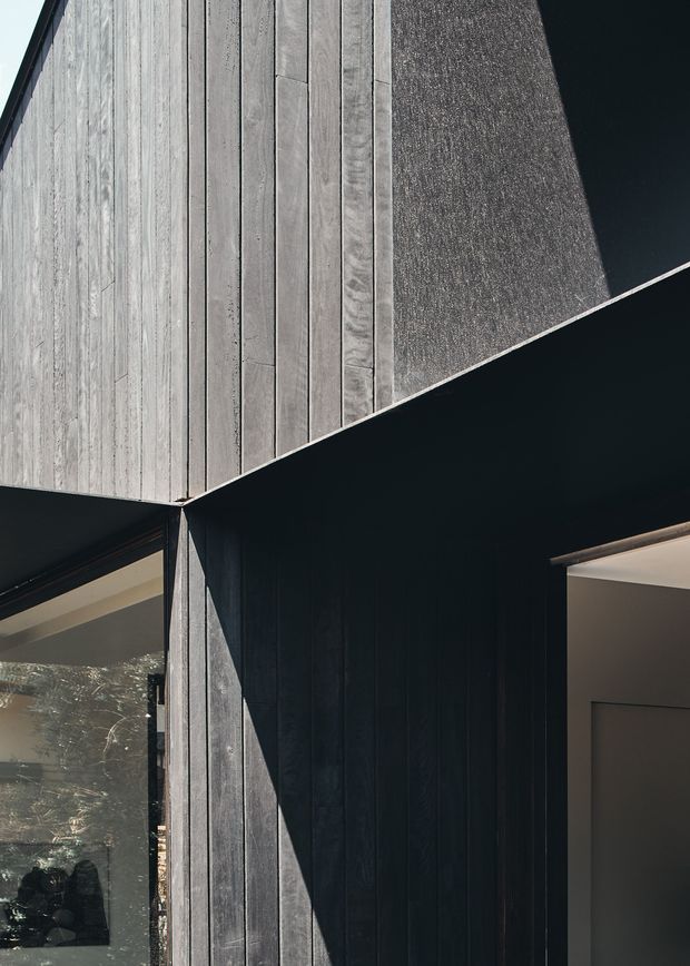 Close-up architectural exterior detail showing a sharply articulated corner clad in vertically laid, weathered timber boards. Deep charcoal-black wall surfaces and deep roof or soffit planes contrast with pale grey timber, while a glazed opening and recessed doorway are visible at the lower edges. Strong sunlight creates dramatic angular shadows across the facade.