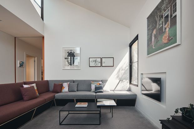 Bright contemporary living room with a high ceiling and clerestory glazing. A large L-shaped sectional sofa spans the corner, facing a slim built-in fireplace and black metal coffee tables. White walls, dark grey carpet, framed artwork, and strong sunlight create a clean, refined interior.