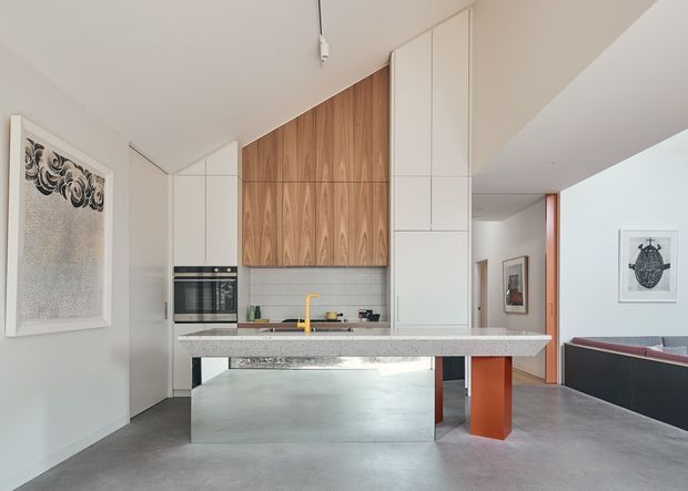 Contemporary open-plan kitchen with a long island bench, integrated white cabinetry, warm timber wall cabinetry, a speckled stone benchtop, and a polished concrete floor. The space opens toward a living area and hallway, with framed artwork and a bold orange structural or joinery element adding contrast.