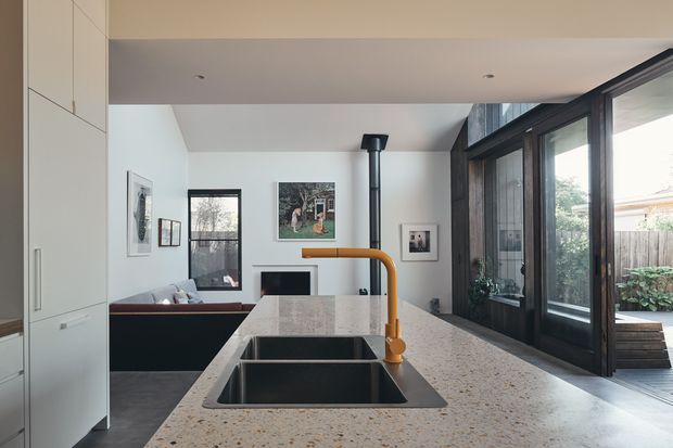 Contemporary open-plan kitchen viewed from the island, with a large speckled stone benchtop, undermount double sink, and distinctive brushed brass gooseneck tap. Full-height dark-framed glazed doors open toward an outdoor deck, while a living area with a fireplace is visible beyond. The space has white built-in cabinetry, dark flooring, and abundant natural light.