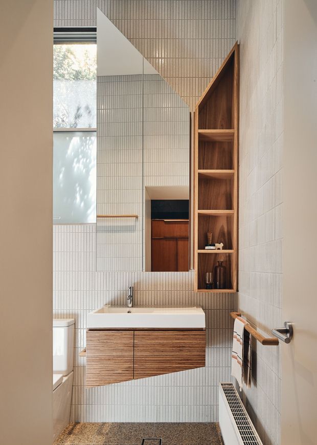 A compact contemporary bathroom viewed through an open doorway, featuring vertically ribbed light tile walls, a wall-mounted timber vanity with white rectangular basin, brushed metal tapware, a mirrored cabinet, open timber shelving, and a toilet partially visible at left. A terrazzo-like floor, tall frosted window, heated towel rail, and linear floor grille complete the practical, refined space.