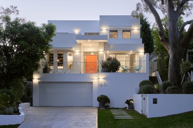 Dusk view of a contemporary multi-level residence with a crisp white rendered facade, flat rooflines, projecting box forms, large windows, and a recessed illuminated entry. The home includes an integrated double garage, upper terrace with outdoor seating, landscaped front garden, paved driveway, and mature trees.