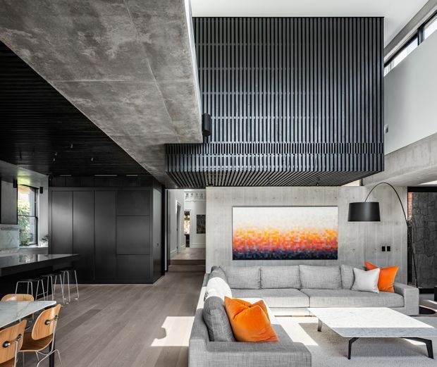 Contemporary open-plan living room with a grey sectional sofa, bright orange cushions, low marble-look coffee tables, and a large abstract artwork. The space features an exposed concrete structural ceiling and walls, dark vertical slatted timber or metal screening around an upper volume, dark built-in cabinetry, black ceiling treatment, and extensive glazing that brings in strong daylight. A compact kitchen or dining area is visible to the left and a hallway extends beyond the living area.